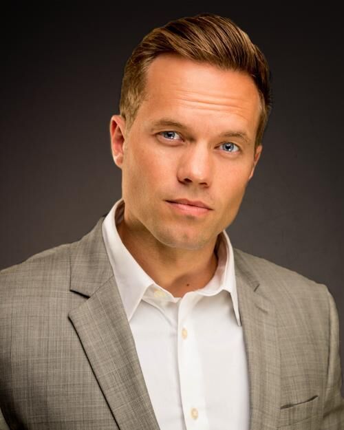 A man in a light gray suit and white shirt posing against a dark background.