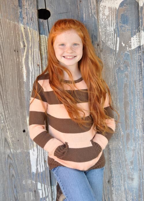 Smiling red-haired girl in a brown striped sweater stands against a wooden wall.