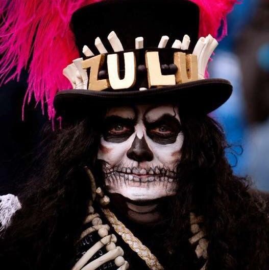 Person in elaborate skull face paint and colorful feathered hat labeled 'ZULU'.