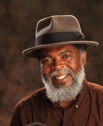 Smiling elderly man with a gray beard wearing a brown hat and shirt.